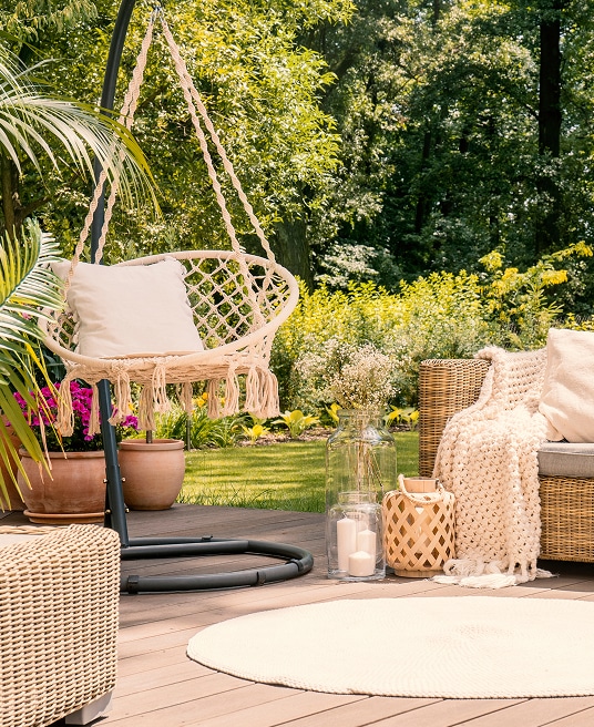 Terrasse bois relaxante avec chaise suspendue macramé, coussin blanc, plaid doux, plantes en pot et décorations bohèmes devant un jardin verdoyant.
