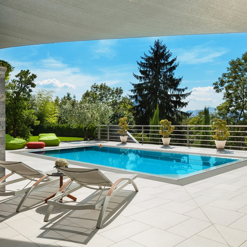 Piscine extérieure moderne, terrasse ensoleillée avec deux transats, et jardin luxuriant. Ciel bleu et arbres, ambiance détente.